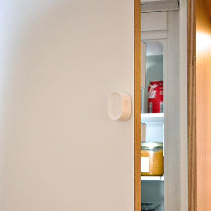Partial view of a refrigerator with a can of Coca-Cola inside, and a Zensor device on it against a neutral wall.